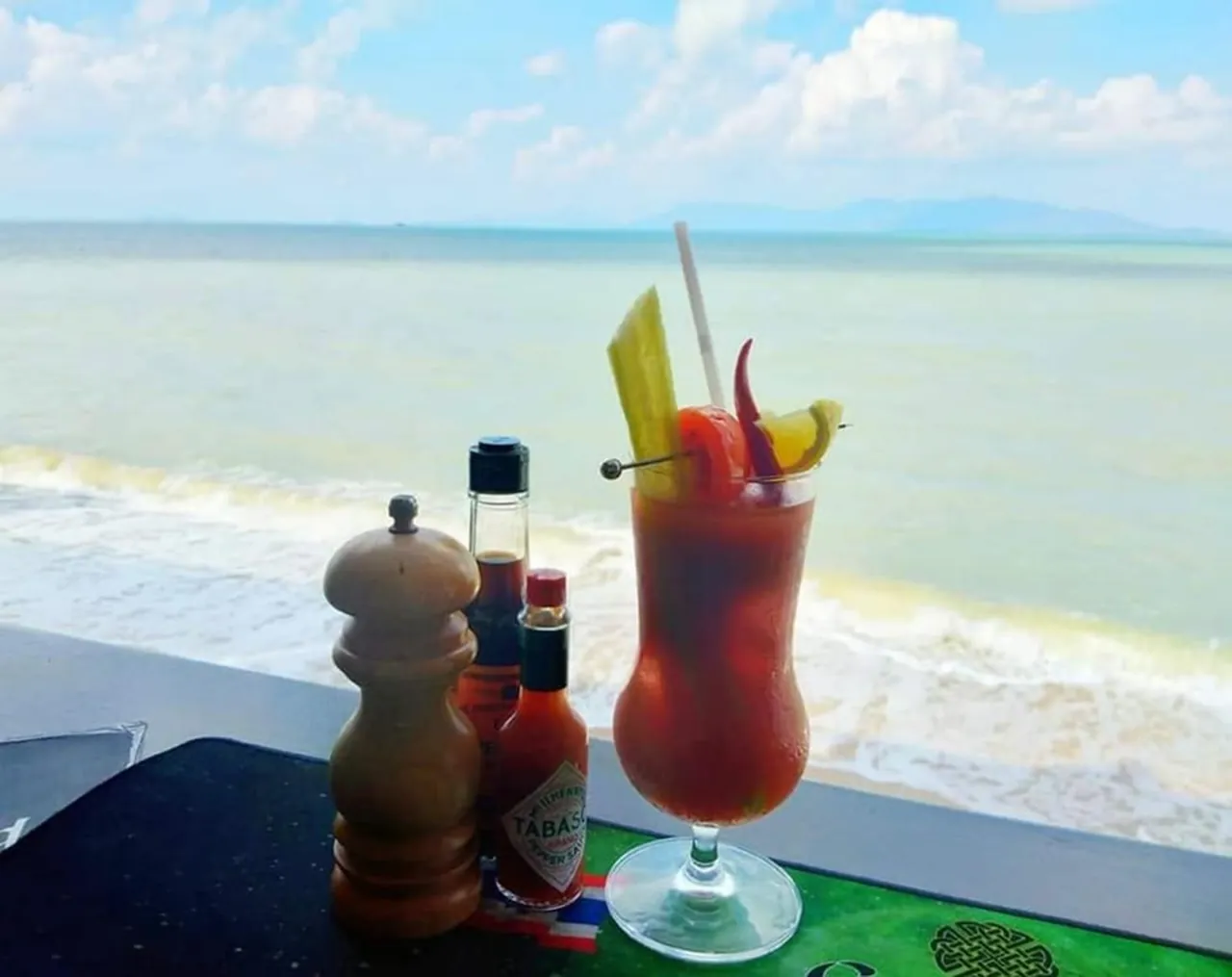 A red cocktail on the bar mat at the sea rail, with the beach and the sea behind