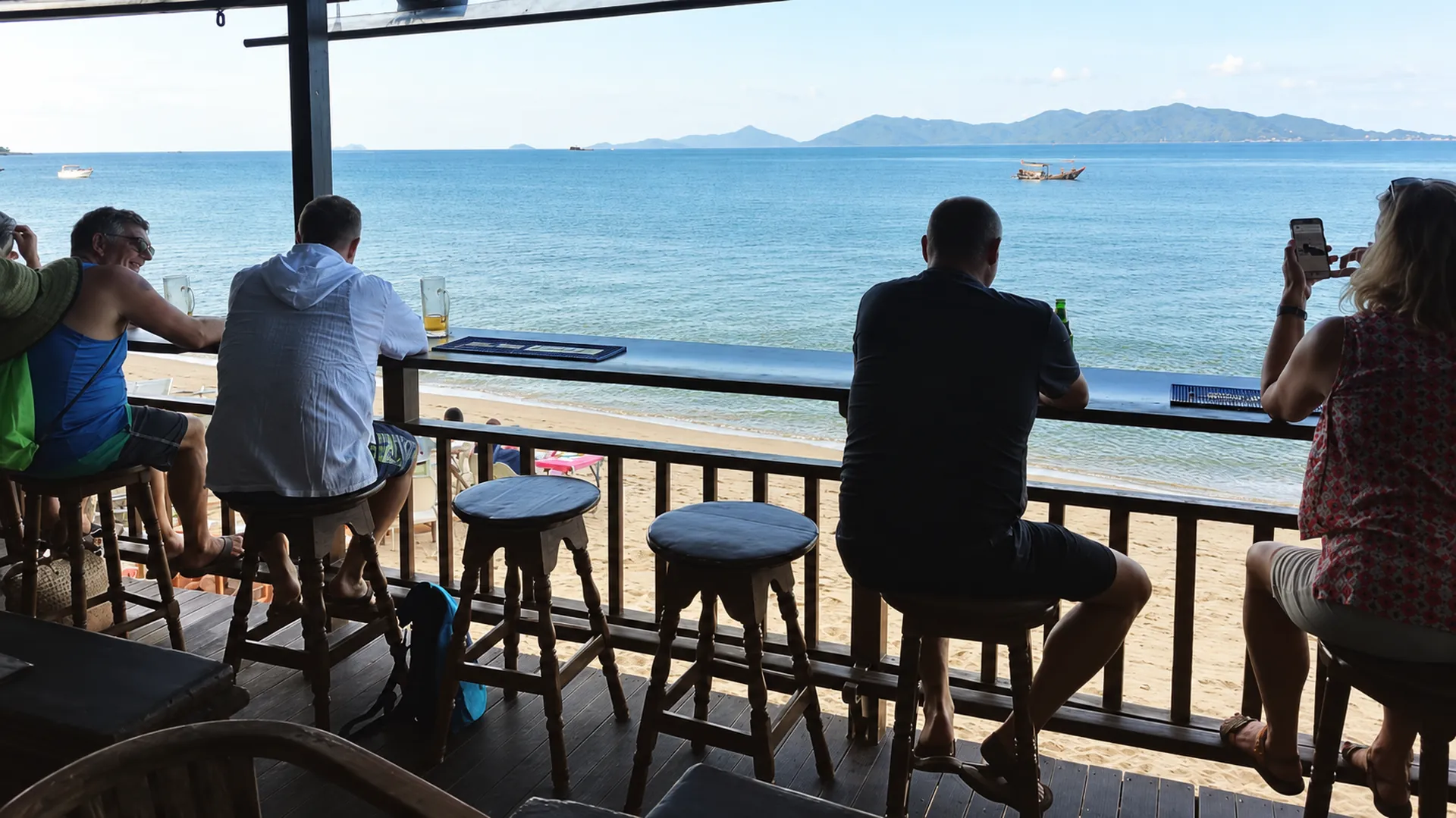 Guests on bar stools at the terrace rail, with the beach and the bay right below